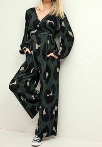 Combinaison avec imprimé léopard vert et noir au design enveloppant, dotée de manches longues, d'une taille à nouer et de poches latérales. Portée avec des baskets noires.