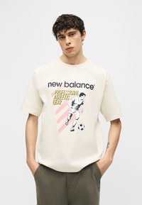 Młody mężczyzna z kręconymi włosami w beżowej koszulce New Balance z grafiką piłkarza i napisem „Professional Athletic Gear”.