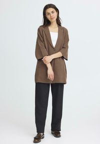 Rudas per didelis švarkas su vienu mygtuku, V formos iškirpte ir plačiomis rankovėmis, derinamas su juodomis plačiomis kelnėmis ir rudais batais.