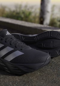 Μαύρα αθλητικά παπούτσια Adidas με γκρι τρεις ρίγες και υφασμένους πάτους που ακουμπούν σε άσφαλτο σε εξωτερικό χώρο.