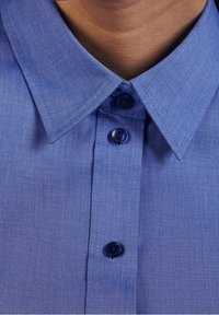 Light blue button-up shirt with a structured collar, featuring a subtle textured pattern and dark blue buttons along the front placket.