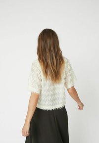 White sheer top with zigzag texture, short sleeves, and frayed edges. Worn over a black skirt, viewed from the back.