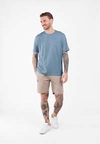 Camisa de manga corta azul claro, pantalones cortos de cargo beige, zapatillas blancas. Tatuajes visibles en los brazos y las piernas. Tejido suave, ajuste casual.