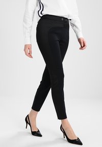 Vrouw die loopt in strakke zwarte broeken, een witte blouse met zijstreepjes en zwarte puntige slingback hakken op een effen achtergrond.
