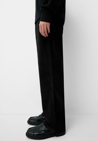 Black corduroy wide-leg trousers with a textured surface, paired with shiny black loafers, captured from the side against a white background.