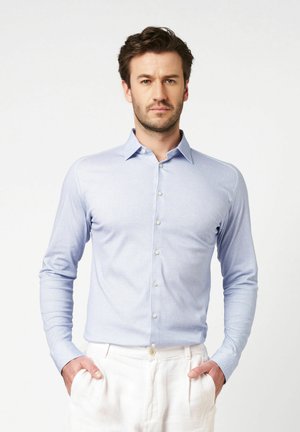 Hellblaues Langarmhemd mit Button-Down-Kragen, feiner Textur und weißen Knöpfen. Dazu helle Leinenhosen getragen.