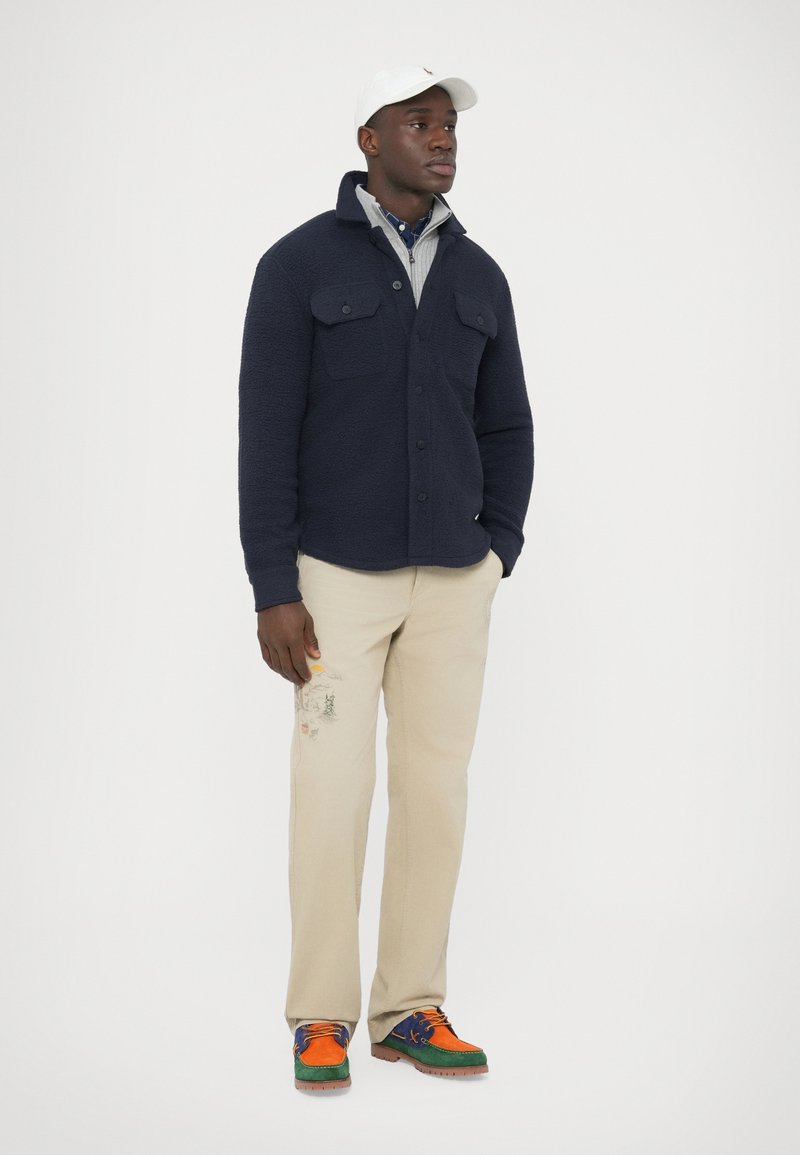 Navy fleece shirt jacket with two chest pockets, beige trousers with embroidered accents, and multicolored sneakers featuring blue, orange, and green.