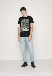 Μαύρο βαμβακερό T-shirt με σχέδιο δύο προσώπων σε γκρι χρώμα στο μπροστινό μέρος. Συνδυασμένο με ανοιχτό μπλε τζιν και μαύρα παπούτσια.