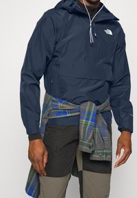 Giacca a vento navy con cappuccio, tasca frontale e cordino regolabile. Flanella a quadri sovrapposta legata in vita. Pantaloni scuri con pannelli testurizzati.