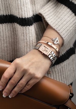 Montre en or rose et argent avec un cadran carré, associée à deux fins bracelets. La main repose sur un sac en cuir marron, portant un pull en tricot beige.