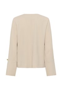 Sieviešu bēša ilgrokšķa žakete ar taisnu muguru, platiem aprocēm un mazu zelta pogas detaļu uz kreisās piedurknes aproces.