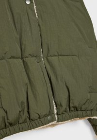 Zbliżenie na oliwkową, pikowaną kurtkę z zatrzaskami i widoczną białą podszewką z polaru wzdłuż krawędzi i dołu.