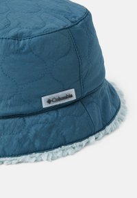 Blå bøttehatt laget av quiltet stoff med et teksturert mønster. Den har en lyseblå faux fur-kant og et Columbia-logo-patch.