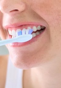 Tandenborstel met blauwe en witte haren, gehouden boven een open mond, die schone, witte tanden en gladde lippen toont, tegen een neutrale achtergrond.