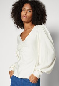 Cream long-sleeve top with a V-neckline, gathered front detail, and voluminous sleeves, paired with blue denim. Soft fabric texture.