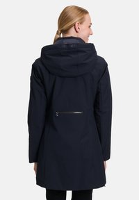 Veste imperméable bleu marine avec capuche, détails en zipper et accents de boutons-pression. Dispose d'un design mi-long et d'une finition lisse et texturée.