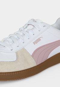 Λευκό και μπεζ αθλητικό παπούτσι Puma με ροζ πλευρική λωρίδα, λευκά κορδόνια και καφέ λαστιχένια σόλα, που δείχνει λεπτομέρειες από μπροστά και πλαϊνά.