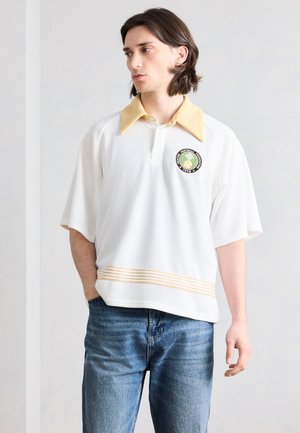 Jeune homme portant une chemise blanche à manches courtes avec col jaune, logo brodé, et un jean bleu, debout devant un mur clair uni.