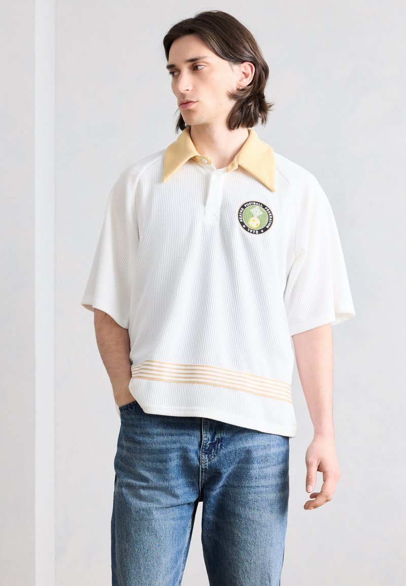 Νεαρός άνδρας με άσπρο κοντομάνικο πουκάμισο με κίτρινο γιακά, κεντητό λογότυπο και μπλε τζιν, στέκεται μπροστά σε απλή ανοιχτόχρωμη επιφάνεια.