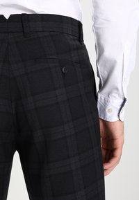 Pantaloni a quadri neri con texture liscia, dotati di chiusura con un solo bottone e tasca posteriore, abbinati a una camicia bianca a maniche lunghe.