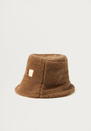 Brown bucket hat made of plush fabric with a textured finish. Features a square metallic logo tag on the side. Rounded, soft shape.