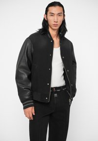 Chaqueta bomber negra con mangas de cuero, puños rayados grises y cierre de botones a presión. Se lleva sobre una camiseta sin mangas blanca y se combina con jeans negros.