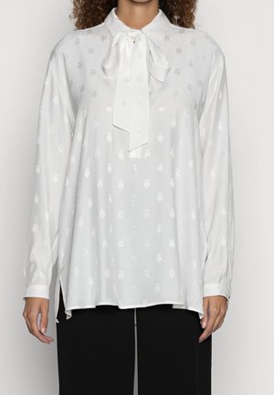 Hvid bluse med lange ærmer og diskrete skinnende logoer, med bundet sløjfe ved kraven, båret over sorte bukser på en person med krøllet hår.