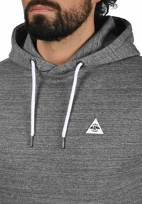 Gray hooded sweatshirt made of textured fabric with white drawstrings. Features a small triangular logo patch on the chest.