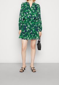 Bloemenjurk met groen bladmotief op een marineblauwe achtergrond, met een V-hals, lange mouwen, ruches aan de zoom, gecombineerd met zwarte sandaaltjes met bandjes.