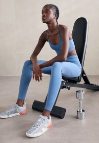 Světle modrý sportovní crop top a legíny, s texturovanými panely, doplněné šedými a růžovými teniskami. Činka leží na podlaze vedle lavičky.