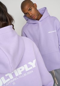 Sudaderas de color lavanda en tejido suave con bolsillo tipo canguro en la parte frontal y logo blanco impreso "MULTIPLY 2024" en la parte trasera.