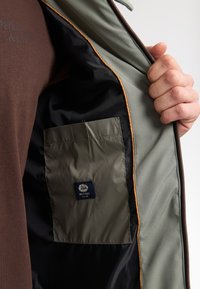 Interiorul jachetei de un verde măsliniu deschis dezvăluie un căptușel negru și un buzunar khaki cu un eticheta de logo albastru. Un material maro este vizibil dedesubtul jachetei.