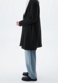 Cappotto di lana nera con un design strutturato, tasche laterali, abbinato a jeans azzurri a gamba dritta e stivali neri con lacci.