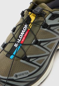 Zelená textúrovaná bežecká obuv s označením GORE-TEX, s čiernymi šnúrkami, žltým a modrým akcentom a hexagónovým vzorom na vrchu.