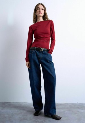 Femme debout contre un mur uni portant un haut rouge à manches longues, un jean bleu foncé ample avec une ceinture noire et des chaussures noires à bout carré.