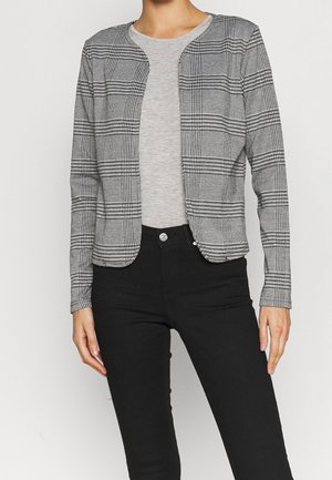 Personne portant un t-shirt gris clair à col rond sous une veste ouverte à carreaux noir et blanc et un pantalon noir ajusté.