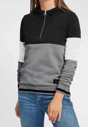 Vrouw draagt een kleurgeblokte hoodie met zwart, wit en grijze secties, halve rits aan de voorkant, gecombineerd met een blauwe spijkerbroek, staand tegen een effen achtergrond.