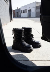 Paar schwarze Plateaustiefel mit Schnürung, auf einem sonnenbeschienenen städtischen Gehsteig neben einer offenen Autotür platziert.
