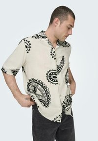 Beige Kurzarmhemd mit schwarzem Paisleymuster, mit Knopfleiste und standardmäßigem Kragen, kombiniert mit dunklen Jeans.