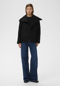Sort uldblandet frakke med en bred, struktureret krave og to knapper, parret med højtaljede, løse blå denimjeans og sorte sko.