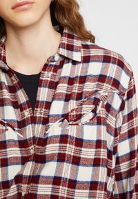 Rotes, blaues und weißes kariertes Flanellhemd mit Knopfleiste, zwei Brusttaschen und strukturiertem Stoff.