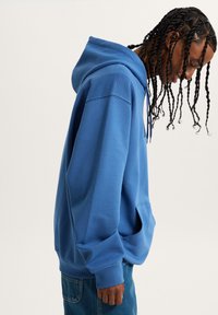 Jeune personne avec des tresses portant un sweat à capuche bleu et un jean, penchée en avant avec les mains relâchées sur le côté, sur un fond uni.