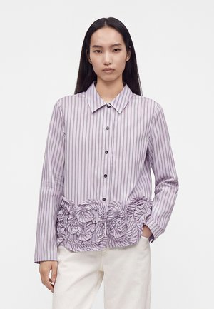 Femme aux longs cheveux noirs portant une chemise rayée violet clair avec boutons et détail froncé à l’ourlet, ainsi qu’un pantalon blanc, debout sur fond blanc.