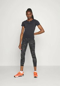 Ženska stoji s roko na boku, oblečena v črno oprijeto majico, črne vojaške legice, bele nogavice in svetlo oranžne superge na enobarvni ozadju.