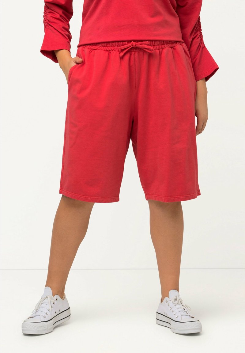 Personne portant un short rouge ample à cordon et un haut à manches longues assorti, debout les mains dans les poches et des baskets blanches.