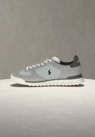 Sive in bele nizke superge z belimi vezalkami, teksturiranimi paneli in črnim Polo logotipom na strani, postavljene na nevtralno ozadje.