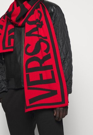 Veste et pantalon matelassés noirs portés avec une écharpe rouge et noire affichant un texte large et audacieux, montrant partiellement le mot « VERSUS ».