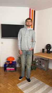 Haut à capuche gris avec une poche kangourou, jeans bleu clair et chaussures noires à enfiler. Se tenant sur un tapis texturé dans un cadre de pièce moderne.