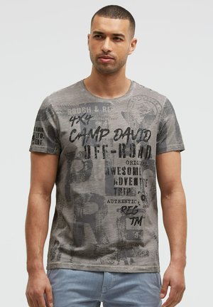 Man with short hair wearing a gray graphic t-shirt with black text and light blue jeans, standing against a plain light background.