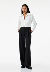 Witte blouse met lange mouwen en knopen, gecombineerd met hoge taille zwarte wijde broek. De outfit heeft een gestroomlijnd ontwerp en een zachte textuur.